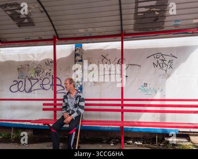 Kiev, Ukraine - 25 juillet 2025 : homme solitaire assis à un arrêt de bus détérioré à Kiev. Ukraine. Attente des transports en commun au milieu des graffitis et ur Banque D'Images