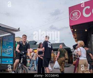 Kiev, Ukraine - 25 juillet 2025 : jeunes adultes socialisant près de bâtiments commerciaux avec un soldat marchant en arrière-plan, illustrant la vie quotidienne i. Banque D'Images