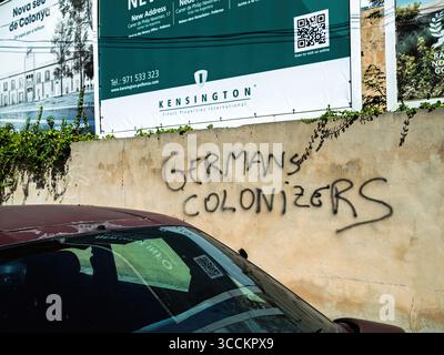 Pollença, Espagne - 202 juillet 2025 : un mur affiche des graffitis lisant les colonisateurs allemands en peinture noire, sous des bannières immobilières faisant la promotion locale Banque D'Images