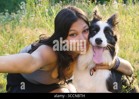 Femme indienne prenant un selfie avec son heureux chien Border Collie dans une prairie ensoleillée, les deux regardant la caméra. Parfait pour les soins des animaux de compagnie, la marche de chien, et co Banque D'Images