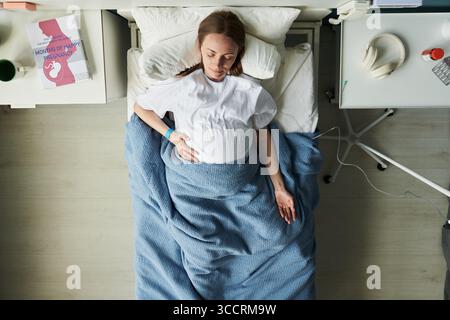 Femme jeune adulte enceinte caucasienne couchée dans un lit d'hôpital reposant la main sur le ventre, équipement médical et livre de maternité visible sur la table de chevet, vue de dessus Banque D'Images