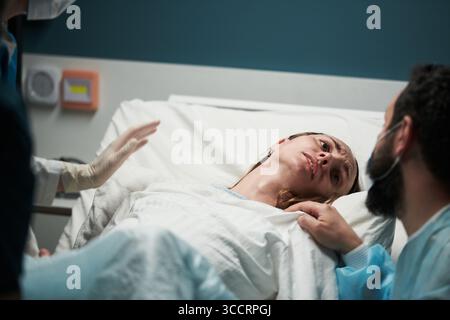 Femme jeune adulte caucasienne couchée dans le lit d'hôpital éprouvant des douleurs de travail, paraissant en détresse tandis que le personnel médical et l'homme caucasien barbu fournissant un soutien pendant l'accouchement Banque D'Images