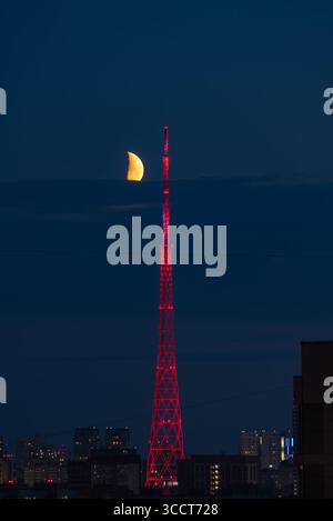 Tour de télévision brillante dans un rouge vif pendant la nuit, accompagnée d'un croissant de lune et d'un paysage urbain époustouflant. La scène capture la beauté de l'architecture urbaine et la tranquillité nocturne Banque D'Images