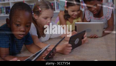 Groupe couché de quatre enfants utilisant des tablettes à écran tactile sur le sol de la bibliothèque en bois, avec des étagères Banque D'Images