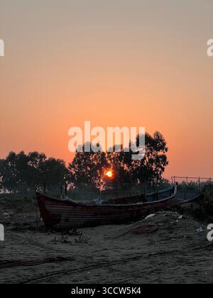 Un lever de soleil à couper le souffle sur la côte nord de l'Iran, avec le soleil doré se levant derrière les arbres et projetant une lumière chaude sur un vieux bateau de pêche en bois. Banque D'Images