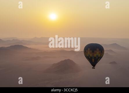 Vue aérienne d'une montgolfière solitaire dérive sereinement au-dessus des dunes ondulées du désert du Namib, baignées par la lueur dorée du soleil levant, Namibie. Banque D'Images