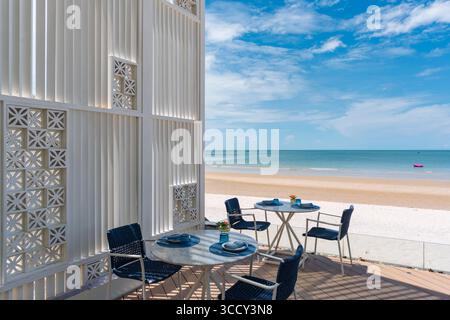 La terrasse élégante du restaurant en bord de mer est dotée d'un mur blanc moderne, de tables en marbre et de chaises bleues les jours ensoleillés, offrant un dîner en plein air relaxant Banque D'Images
