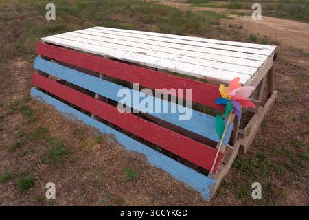 Mobilier d'extérieur créatif et rustique, un simple banc en bois peint en rouge, blanc et bleu, avec un petit jouet. Banque D'Images