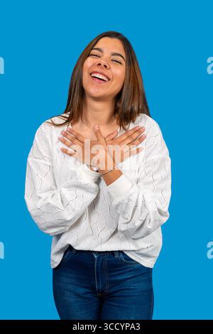 Portrait studio d'une jeune femme reconnaissante tenant les mains sur la poitrine, souriant et exprimant sincèrement gratitude et bonheur Banque D'Images