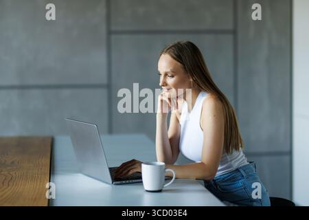 Photo d'une jeune femme attentionnés regardant son ordinateur portable tout en tenant une tasse de café dans la cuisine à la maison. Banque D'Images