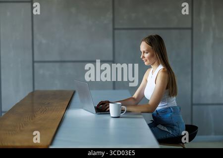 Photo d'une jeune femme attentionnés regardant son ordinateur portable tout en tenant une tasse de café dans la cuisine à la maison. Banque D'Images