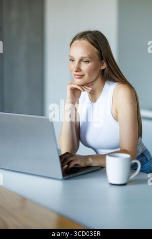 Photo d'une jeune femme attentionnés regardant son ordinateur portable tout en tenant une tasse de café dans la cuisine à la maison. Banque D'Images