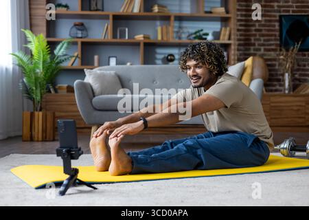 Jeune homme effectuant des étirements sur un tapis de yoga jaune à l'intérieur, accompagné d'un téléphone portable pour l'orientation ou la motivation. La configuration confortable du salon ajoute Banque D'Images