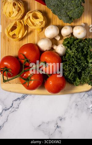 Légumes frais, y compris tomates, champignons, brocoli et chou frisé, ainsi que pâtes alimentaires, sont disposés sur une planche à découper en bois. Banque D'Images