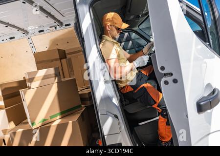 Un livreur est assis dans une camionnette, se préparant pour son itinéraire avec des boîtes chargées à l'intérieur. La lumière du soleil se filtre, mettant en évidence son expression focalisée. Banque D'Images