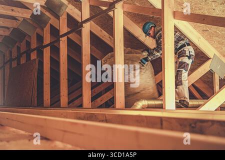 Un travailleur portant un équipement de sécurité place un matériau isolant dans un grenier. Poutres en bois et charpente sont visibles, indiquant des rénovations en cours dans la maison. Banque D'Images