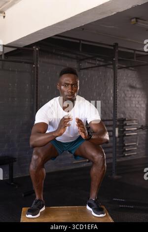 Homme afro-américain accroupissant sur la boîte plyométrique dans la zone de musculation de gymnastique avec support de squat, plaques de musculation Banque D'Images