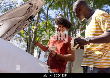Divers amis masculins vérifiant la jauge par la voiture blanche avec le capot ouvert sur l'allée tenant des boissons, chiffon Banque D'Images