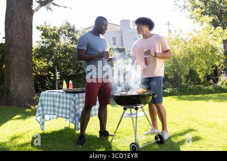 Divers amis masculins barbecue près du gril et table à carreaux avec bouteilles de condiments dans la cour arrière Banque D'Images