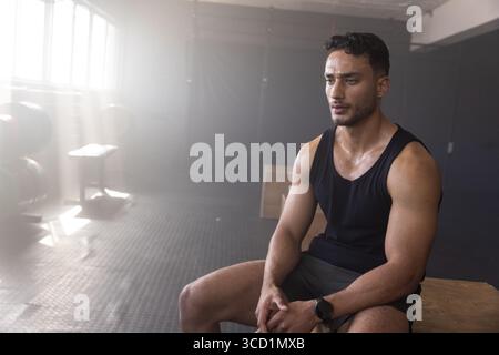 Homme reposant sur la boîte plyométrique dans la salle de gym en débardeur noir et short athlétique, espace copie Banque D'Images