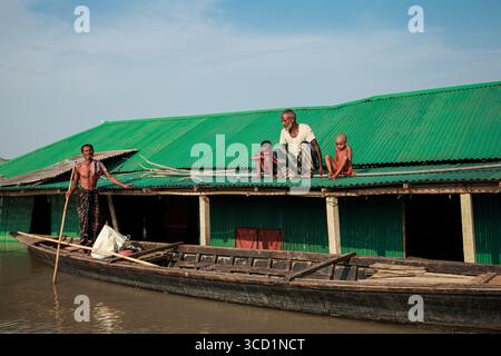 Kurigram, Bangladesh - 21 juin 2022 : vue d'une famille sur un toit vert ondulé, avec un homme debout dans un bateau, au milieu des eaux de crue reflétant le Banque D'Images