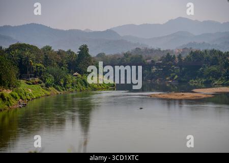 Vue paisible sur la rivière Nam Khan et le pont en bois au milieu d'une végétation luxuriante et de paysages de montagne à Luang Prabang, Laos Banque D'Images