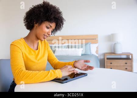 Femme afro-américaine portant le dessus de moutarde en utilisant la tablette à la table dans la chambre à coucher par le lit, espace de copie Banque D'Images