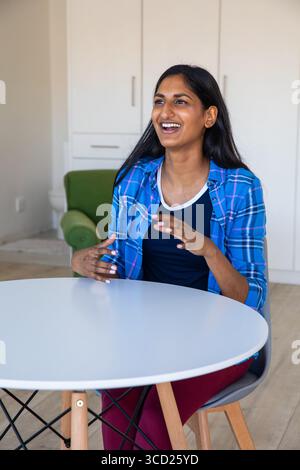 Femme indienne assise à une table blanche ronde dans le coin salon avec fauteuil vert en utilisant des gestes de la main Banque D'Images