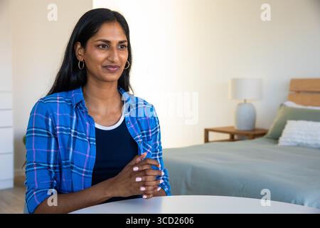 Femme indienne assise portant chemise à carreaux serrant les mains dans la chambre à coucher à la table à côté de la lampe, espace de copie Banque D'Images