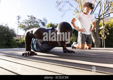 Entraîneur masculin diversifié guidant le stagiaire effectuant des pompes sur le pont en bois à l'extérieur avec smartwatch Banque D'Images