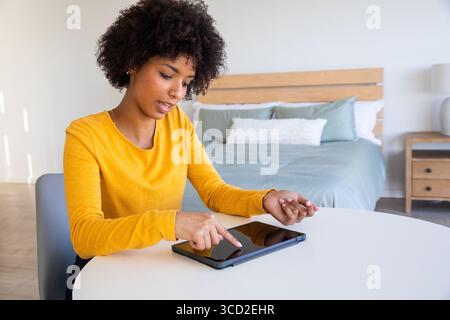 Femme afro-américaine portant le dessus jaune moutarde penché à la table dans la chambre à coucher à l'aide d'une tablette noire Banque D'Images