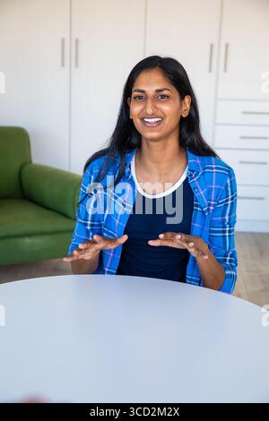Femme indienne faisant des gestes et souriant à table dans un salon avec armoires encastrées et fauteuil vert Banque D'Images