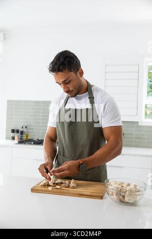 Homme dans le tablier d'olive hachant des champignons sur une planche à découper en bois avec bol en verre à l'îlot de cuisine Banque D'Images