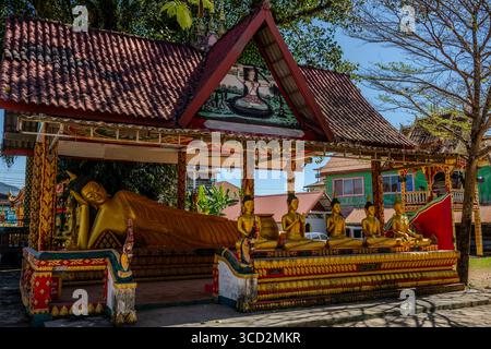 Bouddha couché doré et statues assises au temple bouddhiste de Vang Vieng, Laos avec architecture traditionnelle ornée et détail de toit peint Banque D'Images