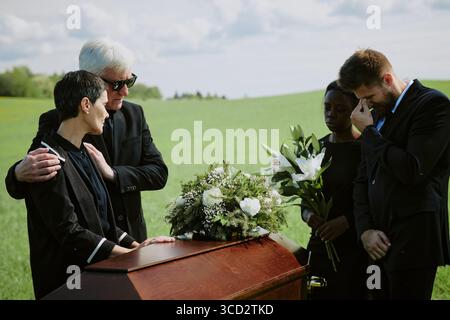 Homme âgé caucasien réconfortant femme caucasienne d'âge moyen debout à côté du cercueil à l'extérieur, jeune femme adulte noire tenant des lis, jeune homme adulte caucasien essuyant les yeux Banque D'Images