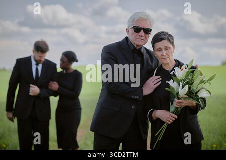 Homme caucasien senior portant des lunettes de soleil réconfortant femme caucasienne d'âge moyen tenant un bouquet de lis tout en se tenant debout à l'extérieur, femme noire et homme caucasien embrassant en arrière-plan Banque D'Images