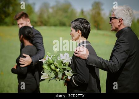 Femme caucasienne d'âge moyen tenant un bouquet de lis debout à côté d'un homme caucasien senior la réconfortant, jeune femme noire et jeune homme caucasien marchant devant dans un cadre extérieur herbeux Banque D'Images