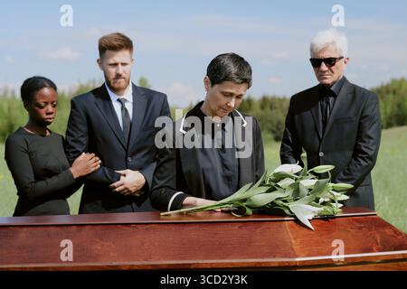 Femme caucasienne d'âge moyen tenant des lis debout près d'un cercueil en bois à l'extérieur, femme noire et homme caucasien debout ensemble, homme caucasien senior portant des lunettes de soleil debout à proximité Banque D'Images