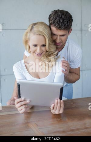 Jeune couple lisant des informations sur une tablette avec la belle femme blonde assise à une table avec son mari lisant par-dessus son épaule Banque D'Images
