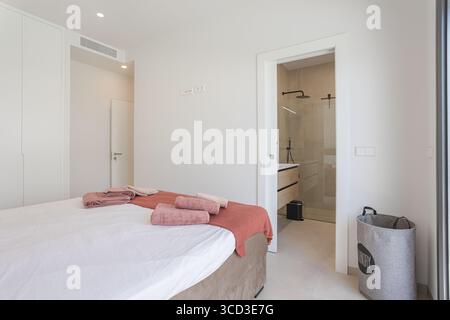 Chambre lumineuse avec murs blancs, serviettes roses douces et accès direct à une salle de bains moderne avec douche à l'italienne. Banque D'Images
