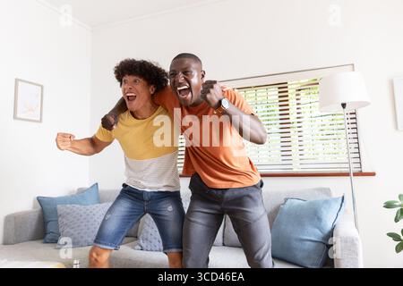 Divers amis masculins acclamant tout en se tenant debout sur un canapé gris clair sous des stores en bois dans le salon Banque D'Images