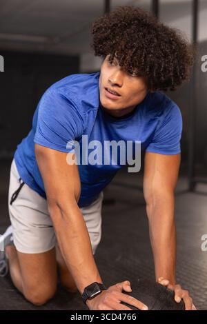 Homme afro-américain adulte agenouillé sur le sol de la salle de sport saisissant le medicine ball texturé portant smartwatch Banque D'Images