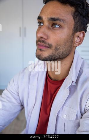 Homme asiatique regardant vers le haut assis dans la chambre près du placard blanc portant un t-shirt rouge, chemise rayée Banque D'Images