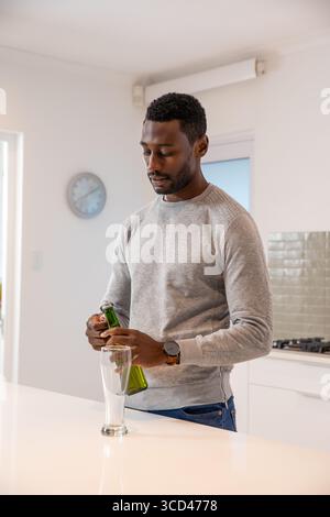 Homme afro-américain versant de la bière de la bouteille verte dans le grand verre transparent sur l'île blanche de la cuisine Banque D'Images