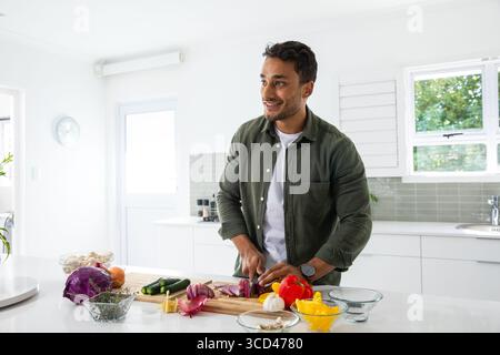 Homme asiatique debout au comptoir blanc dans la cuisine moderne hachant l'oignon rouge avec le couteau du chef Banque D'Images