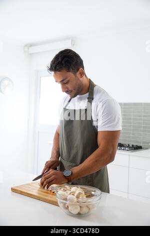 Homme hachant des champignons sur une planche à découper en bois au comptoir de cuisine à la maison portant un tablier vert olive Banque D'Images
