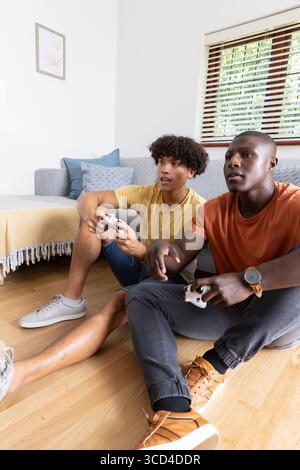 Divers amis masculins assis sur le sol dans le salon tenant deux manettes de jeu par des coussins de canapé Banque D'Images