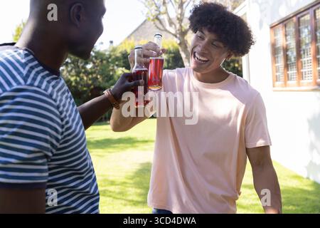 Divers amis masculins clinquant des bouteilles en verre clair remplies de boisson rouge sur la pelouse de l'arrière-cour Banque D'Images