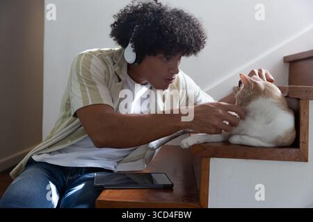 Homme afro-américain accroupi dans les escaliers avec des écouteurs blancs caressant chat à côté de la tablette à la maison Banque D'Images