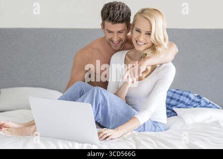 Attrayant, joyeux et souriant jeune couple se relaxant au lit dans leurs vêtements de nuit en utilisant un ordinateur portable pendant qu'ils surfent sur Internet Banque D'Images